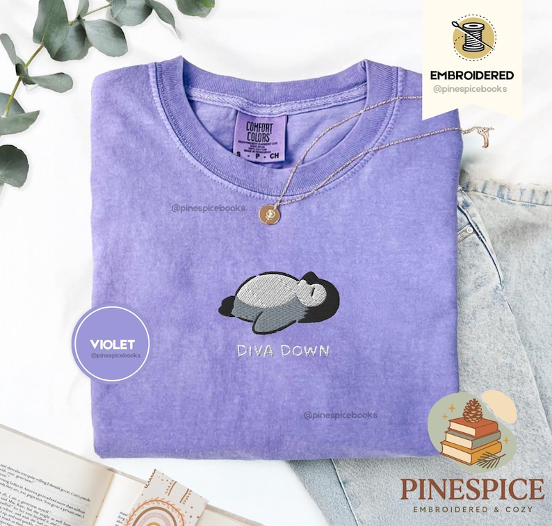 May include: A violet Comfort Colors t-shirt with an embroidered design of a sleeping penguin and the words "DIVA DOWN." A gold necklace and a "VIOLET" circle are also visible. The shirt is folded on a light surface.