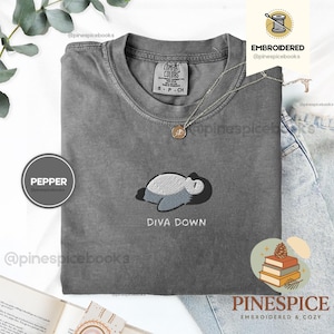 May include: A grey Comfort Colors t-shirt featuring an embroidered design of a sleeping creature and the words "DIVA DOWN." The shirt has a crew neck and short sleeves. A silver necklace with a pendant is also visible.