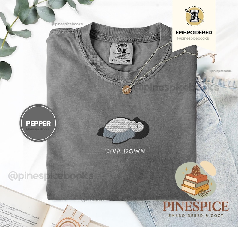 May include: A grey Comfort Colors t-shirt featuring an embroidered design of a sleeping creature and the words "DIVA DOWN." The shirt has a crew neck and short sleeves. A silver necklace with a pendant is also visible.