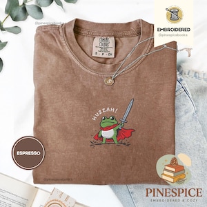 May include: Espresso-coloured t-shirt featuring an embroidered frog warrior design. The frog is green and white, wearing a red cape and holding a sword. The word "HUZZAH!" is above the frog. The shirt is a Comfort Colors brand.
