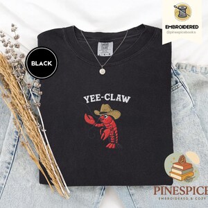 May include: Black sweatshirt with the embroidered phrase "YEE-CLAW" above a red lobster wearing a cowboy hat. The sweatshirt is a Comfort Colors brand. The word "BLACK" is in a black circle on the upper left.