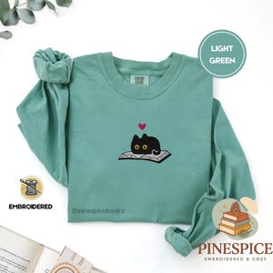 Puede incluir: Una sudadera de manga larga y cuello redondo en color verde claro con un bordado de un gato negro de ojos amarillos sobre un libro abierto, coronado por un pequeño corazón rosa. La etiqueta Comfort Colors aparece en el cuello interior.