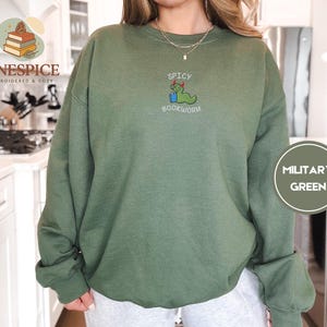 Puede incluir: Sudadera verde militar con la frase bordada "Spicy Bookworm" y un gusano de libro de dibujos animados con gafas y birrete. La sudadera es de color liso y tiene mangas largas.