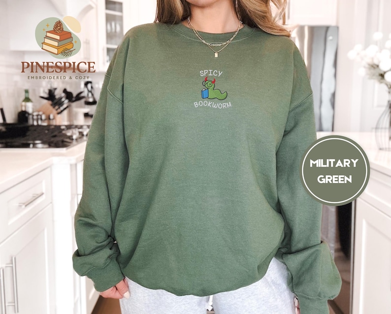 Puede incluir: Sudadera verde militar con la frase bordada "Spicy Bookworm" y un gusano de libro de dibujos animados con gafas y birrete. La sudadera es de color liso y tiene mangas largas.