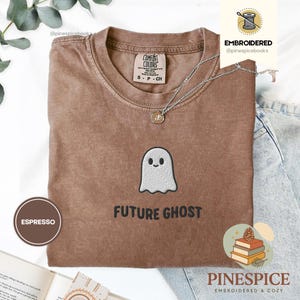 May include: Espresso-colored t-shirt with a crew neck and short sleeves. The shirt features an embroidered ghost design and the text "FUTURE GHOST" in black. A silver necklace with a pendant is visible. The shirt is from Comfort Colors.