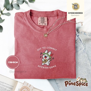 Può includere: Una t-shirt a maniche corte color cremisi con un ricamo di un coniglio bianco che indossa un cappello verde e suona il liuto. Il testo sopra e sotto il coniglio recita "OFF TO COMMIT SHENANIGANS" in filo bianco. Una collana argentata poggia sul colletto.