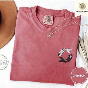 May include: Crimson red t-shirt with an embroidered design of two black and white fish. The shirt is made of 100% cotton and is pre-shrunk. The text "COMFORT COLORS" and "S-P-CH" is printed on the tag.