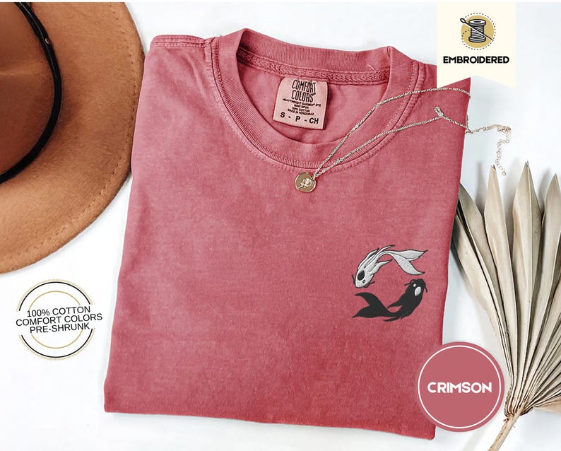 May include: Crimson red t-shirt with an embroidered design of two black and white fish. The shirt is made of 100% cotton and is pre-shrunk. The text "COMFORT COLORS" and "S-P-CH" is printed on the tag.