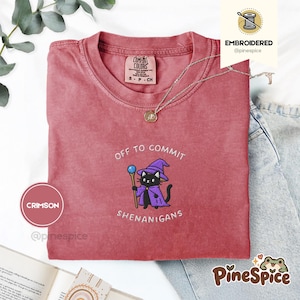 Puede incluir: Una camiseta carmesí con un bordado de un gato negro con sombrero y capa de mago morados, sosteniendo un bastón con una esfera azul. El texto dice OFF TO COMMIT SHENANIGANS. Etiqueta de la marca Comfort Colors en el cuello.
