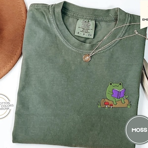 May include: A green t-shirt with a frog reading a book embroidered on the front. The shirt is 100% cotton, comfort colors, and pre-shrunk. The text "COMFORT COLORS" and "S - P - CH" is on a tag attached to the shirt.