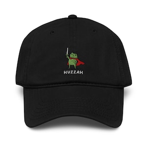 Op de afbeelding: Zwarte baseball cap met een geborduurd ontwerp van een groene kikker met een rode cape en een zwaard. Het woord "Huzzah" is eronder geborduurd. De cap heeft een gebogen klep en een verstelbare band.