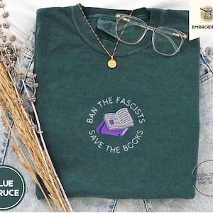 Könnte beinhalten: Ein gefaltetes blaues Fichten-T-Shirt mit der gestickten Aufschrift "BAN THE FASCISTS SAVE THE BOOKS" und einem Bild eines offenen Buches. Das Shirt besteht aus 100 % Baumwolle und hat eine vorgeschrumpfte Komfortpassform. Eine goldene Halskette und eine klare Brille liegen auf dem Shirt.