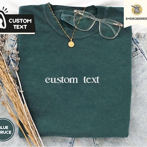 Op de afbeelding: Een donkergroen t-shirt met de woorden "custom text" in het wit geborduurd. Een gouden ketting met hanger en een bril met helder montuur liggen op het shirt. Het shirt ligt op een denim oppervlak met gedroogde bloemen.