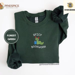 Puede incluir: Sudadera verde bosque con dise&ntilde;o bordado "Spicy Bookworm". El dise&ntilde;o presenta un gusano verde con gafas sosteniendo un libro azul. La sudadera tiene cuello redondo y mangas largas.