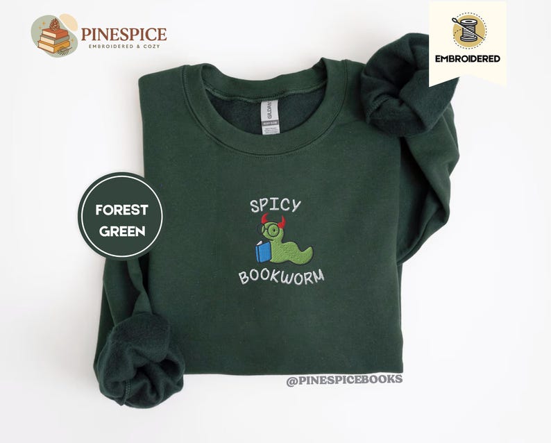 Puede incluir: Sudadera verde bosque con dise&ntilde;o bordado "Spicy Bookworm". El dise&ntilde;o presenta un gusano verde con gafas sosteniendo un libro azul. La sudadera tiene cuello redondo y mangas largas.