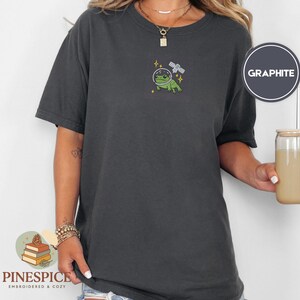 Pu&ograve; includere: T-shirt color grafite con un design ricamato di una rana verde con un casco spaziale e un satellite. La maglietta ha maniche corte e girocollo. Il marchio "Pinespice" &egrave; in basso a sinistra.