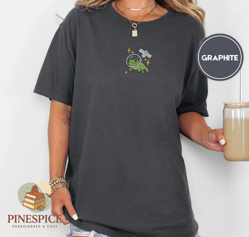 Pu&ograve; includere: T-shirt color grafite con un design ricamato di una rana verde con un casco spaziale e un satellite. La maglietta ha maniche corte e girocollo. Il marchio "Pinespice" &egrave; in basso a sinistra.