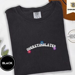 May include: Black t-shirt with the word "OVERSTIMULATED" embroidered in white, with small flower accents in blue, pink, and purple. The shirt is made of 100% cotton and is pre-shrunk. The shirt is a Comfort Colors brand.