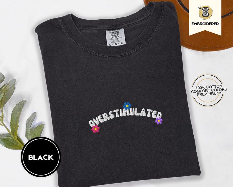 May include: Black t-shirt with the word "OVERSTIMULATED" embroidered in white, with small flower accents in blue, pink, and purple. The shirt is made of 100% cotton and is pre-shrunk. The shirt is a Comfort Colors brand.