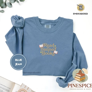 Op de afbeelding: Een spijkerblauw sweatshirt met lange mouwen en een ronde hals, voorzien van geborduurde gele tekst met de woorden Read Banned Books, geaccentueerd door kleine witte iconen van open boeken en vierpuntige sterren. Geribbelde kraag en boorden.