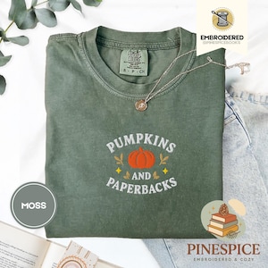 May include: A moss green t-shirt with the embroidered text "PUMPKINS AND PAPERBACKS" and a pumpkin graphic. The shirt has a crew neck and short sleeves. A silver necklace with a pendant is draped across the shirt. The shirt is folded on a light blue denim surface.