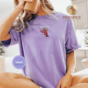 May include: A violet t-shirt with a small embroidered design of a lobster wearing a cowboy hat and the text "Yee-Claw". The shirt has short sleeves and a relaxed fit. The shirt is part of the "PineSpice" collection.