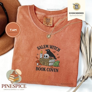Könnte beinhalten: Ein orangefarbenes Comfort Colors T-Shirt mit dem gestickten Text "Salem Witch Book Coven". Das Design umfasst einen Raben, einen Totenkopf, Kerzen, Pilze und einen Frosch. Eine goldene Halskette und ein brauner Hut sind ebenfalls sichtbar.
