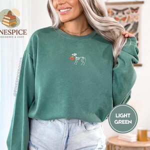 Puede incluir: Sudadera verde claro con un dise&ntilde;o de perro esqueleto sosteniendo una calabaza. La sudadera tiene mangas largas y cuello redondo. El texto "PINESPICE EMBROIDERED & COZY" es visible en el lado izquierdo de la imagen.