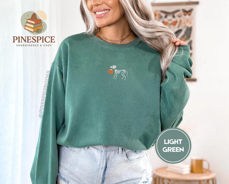Puede incluir: Sudadera verde claro con un dise&ntilde;o de perro esqueleto sosteniendo una calabaza. La sudadera tiene mangas largas y cuello redondo. El texto "PINESPICE EMBROIDERED & COZY" es visible en el lado izquierdo de la imagen.