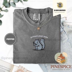 Puede incluir: Una camiseta gris carbón con un bordado de una ardilla sosteniendo un cuchillo pequeño, con el texto "protect your nuts" arqueado sobre la ardilla en hilo blanco. La camiseta tiene un cuello redondo y una etiqueta de marca visible en el cuello.