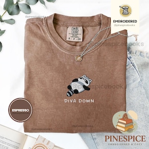 Puede incluir: Una camiseta marrón de manga corta con cuello redondo que presenta un bordado de un mapache tumbado boca arriba. Debajo del mapache, el texto DIVA DOWN aparece en letras blancas. La camiseta incluye una etiqueta de la marca Comfort Colors.