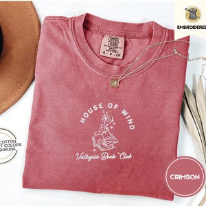 以下が含まれることがあります： ワインレッドのTシャツに、白い刺繍で花がついた本と「House of Wind Valkyrie Book Club」の文字がデザインされています。