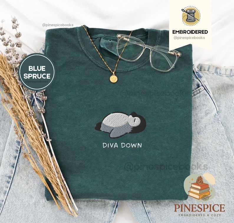 May include: A teal t-shirt featuring an embroidered penguin design with the text "DIVA DOWN". The shirt is styled with a gold necklace and clear-framed glasses. The words "BLUE SPRUCE" are in a circle on the left.