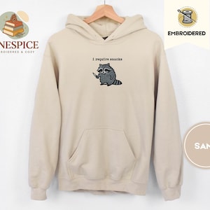 May include: Sand-colored hoodie with a kangaroo pocket and a drawstring hood. The embroidered design features a raccoon holding a knife, with the text "I require snacks" above it. The hoodie is displayed on a wooden hanger.