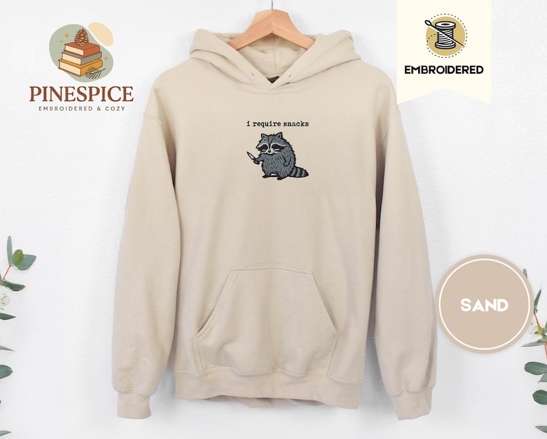 May include: Sand-colored hoodie with a kangaroo pocket and a drawstring hood. The embroidered design features a raccoon holding a knife, with the text "I require snacks" above it. The hoodie is displayed on a wooden hanger.