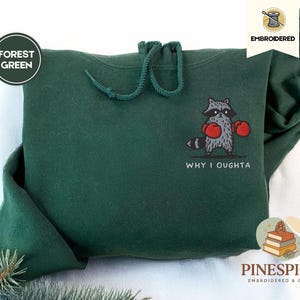 May include: Forest green hoodie with a drawstring hood. The front features an embroidered raccoon wearing red boxing gloves, with the text "WHY I OUGHTA" below. The hoodie is from Pinespice, embroidered and cosy.