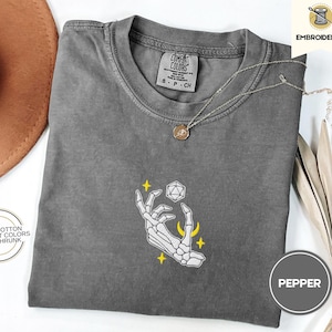 May include: A gray t-shirt with a white embroidered design of a skeletal hand holding a 20-sided die with stars and crescent moons around it. The text "COMFORT COLORS" and "8 - P - CH" is visible on the shirt.