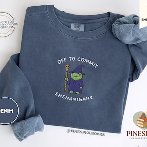 Może przedstawiać: Niebieska dżinsowa bluza z haftowanym napisem "Off to Commit Shenanigans" i rysunkiem żaby-czarodzieja. Bluza ma okrągły dekolt i długie rękawy. Widoczny jest również napis "100% Cotton Comfort Colors Pre-Shrunk".