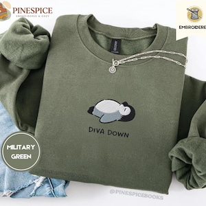 May include: Military green sweatshirt with an embroidered design of a sleeping penguin and the words "DIVA DOWN". The sweatshirt has a crew neck and long sleeves. A silver necklace with a small pendant is on the sweatshirt. The sweatshirt is folded on top of a pair of blue jeans.