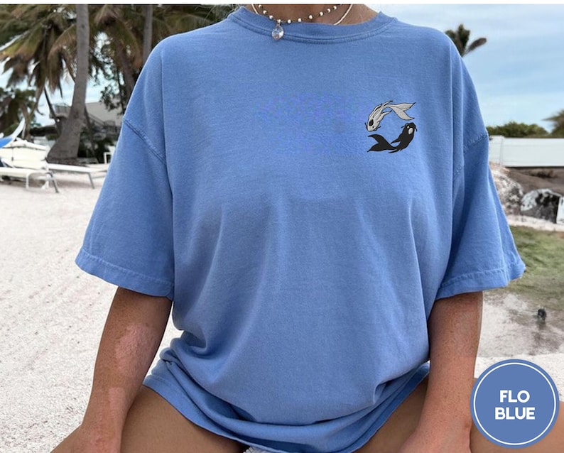 May include: A light blue t-shirt with a graphic of two fish swimming in a circle. The fish are black and white. The shirt is made of a soft, comfortable material.