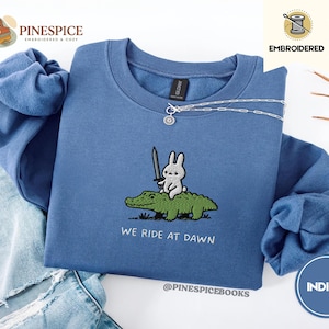 May include: Indigo blue sweatshirt with an embroidered design of a bunny holding a sword riding a green alligator. The text "WE RIDE AT DAWN" is below the design. The brand name "PINESPICE" is visible.