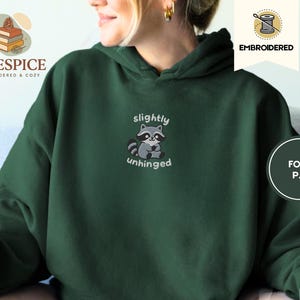 Pu&ograve; includere: Felpa con cappuccio verde scuro con un design ricamato di procione. Il procione &egrave; grigio e nero, con le parole "slightly unhinged" sopra e sotto. La felpa ha un cappuccio con coulisse e una tasca a marsupio.