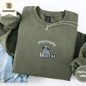 Könnte beinhalten: Militärgrünes Sweatshirt mit dem gestickten Schriftzug "stay trashy" und einem Waschbären, der eine Kaffeetasse hält. Das Sweatshirt liegt gefaltet auf einer Jeans.