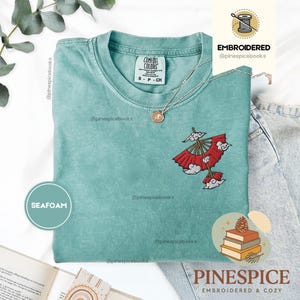 Può includere: Una t-shirt a maniche corte color verde schiuma di mare con un piccolo ricamo sul petto sinistro raffigurante un ombrello rosso in stile giapponese circondato da nuvole bianche. La maglia presenta un girocollo e un'etichetta Comfort Colors interna.