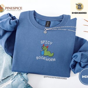 Puede incluir: Sudadera azul &iacute;ndigo con la frase bordada "Spicy Bookworm" y un gusano de libro de dibujos animados con gafas y un libro. La sudadera tiene cuello redondo y mangas largas. Una cadena de plata est&aacute; drapeada en la parte delantera.