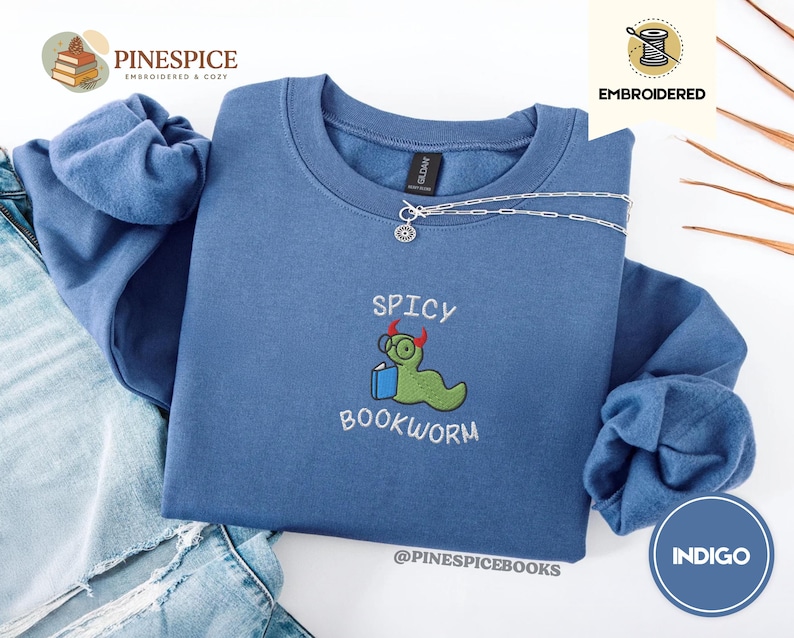 Puede incluir: Sudadera azul &iacute;ndigo con la frase bordada "Spicy Bookworm" y un gusano de libro de dibujos animados con gafas y un libro. La sudadera tiene cuello redondo y mangas largas. Una cadena de plata est&aacute; drapeada en la parte delantera.