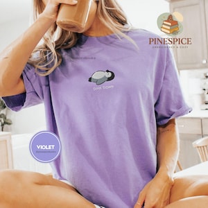 May include: A lilac t-shirt featuring an embroidered penguin and the text "DIVA DOWN". The shirt also has the word "VIOLET" in a circle. The brand name "PINESPICE" is also visible.