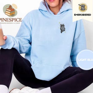 K&ouml;nnte beinhalten: Hellblauer Hoodie mit gesticktem Waschb&auml;ren-Design. Der Hoodie zeigt die Schriftz&uuml;ge "EMBROIDERED" und "LIGHT BLUE". Der Markenname "PINESPICE" ist ebenfalls sichtbar. Ein gem&uuml;tliches und bequemes Kleidungsst&uuml;ck.