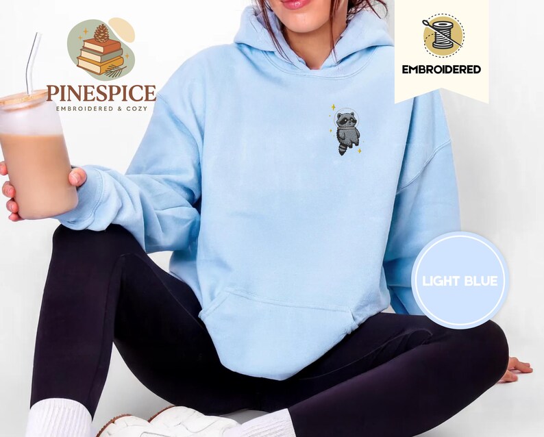 K&ouml;nnte beinhalten: Hellblauer Hoodie mit gesticktem Waschb&auml;ren-Design. Der Hoodie zeigt die Schriftz&uuml;ge "EMBROIDERED" und "LIGHT BLUE". Der Markenname "PINESPICE" ist ebenfalls sichtbar. Ein gem&uuml;tliches und bequemes Kleidungsst&uuml;ck.