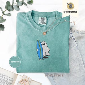 Op de afbeelding: Comfort Colors t-shirt in zeegroen met een geborduurde geest die een blauw en wit surfboard vasthoudt. Het shirt heeft een ronde hals en korte mouwen. Een zilveren ketting met een hanger is over het shirt gedrapeerd. Het woord "EMBROIDERED" staat rechtsboven.